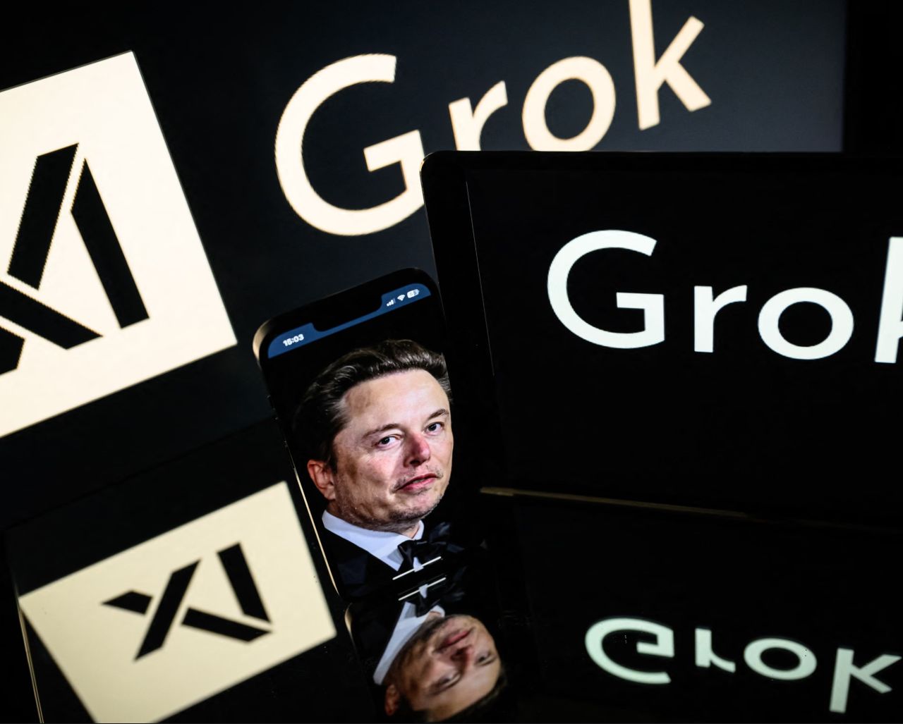 Logo di Grok e di xAI ripetuti su più schermi, con Elon Musk visibile sul display di uno smartphone